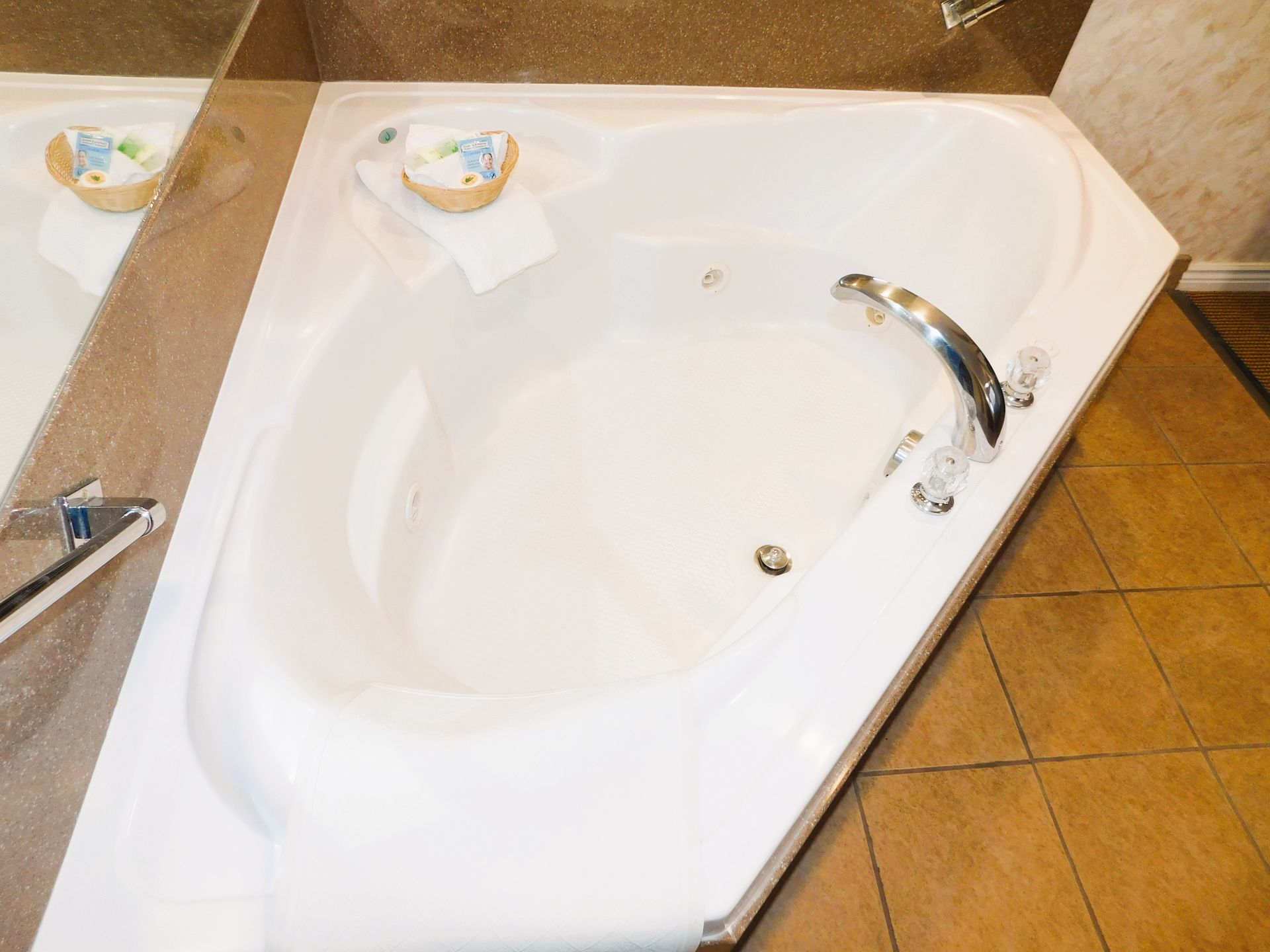 White corner jacuzzi bathtub with chrome fixtures, tiled floor, and a small wooden tray.