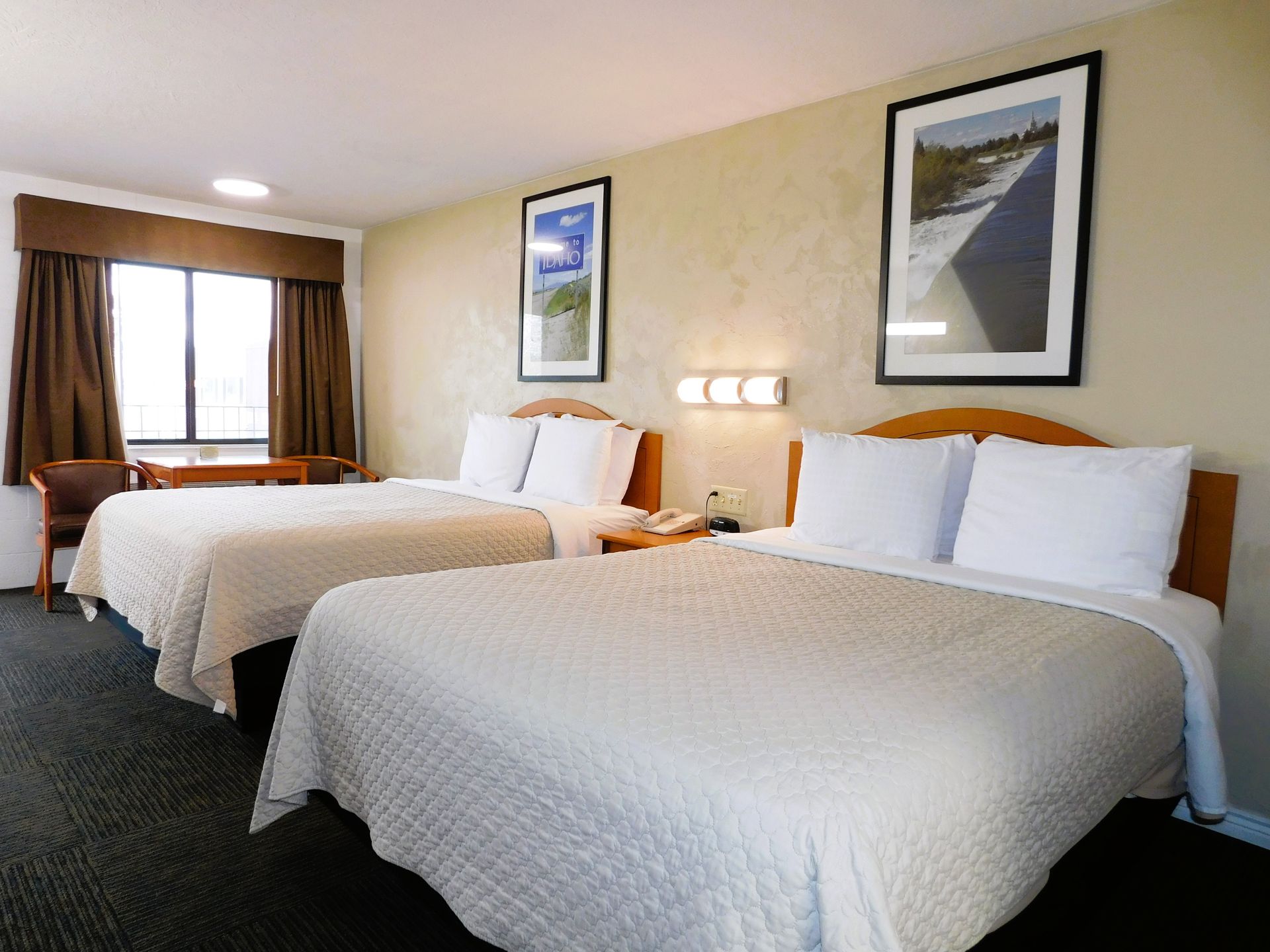Two beds in a motel room, with white bedding, brown curtains, and nature prints on the wall.