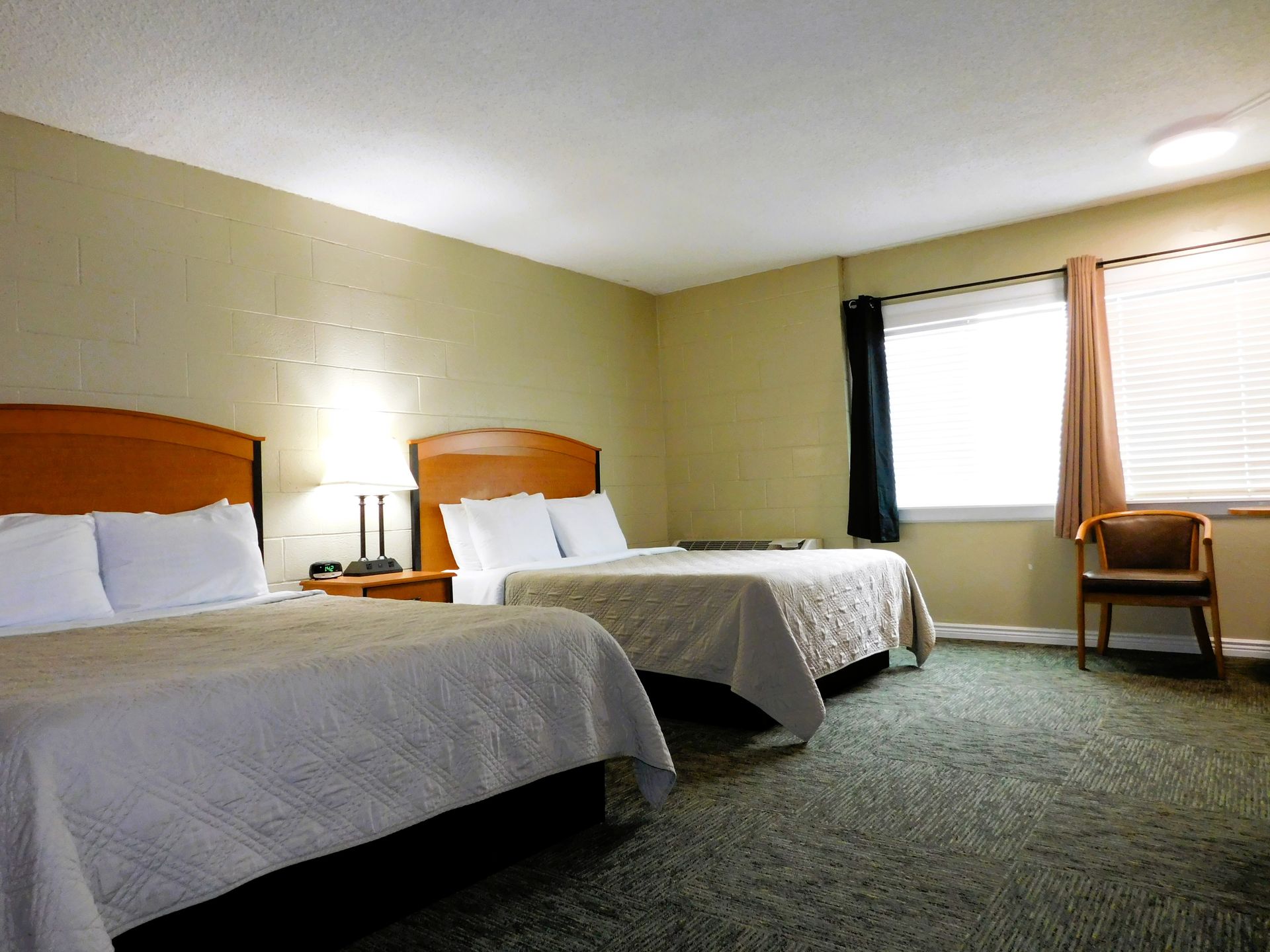 A hotel room with two beds, a window, and a chair; neutral tones and carpet.