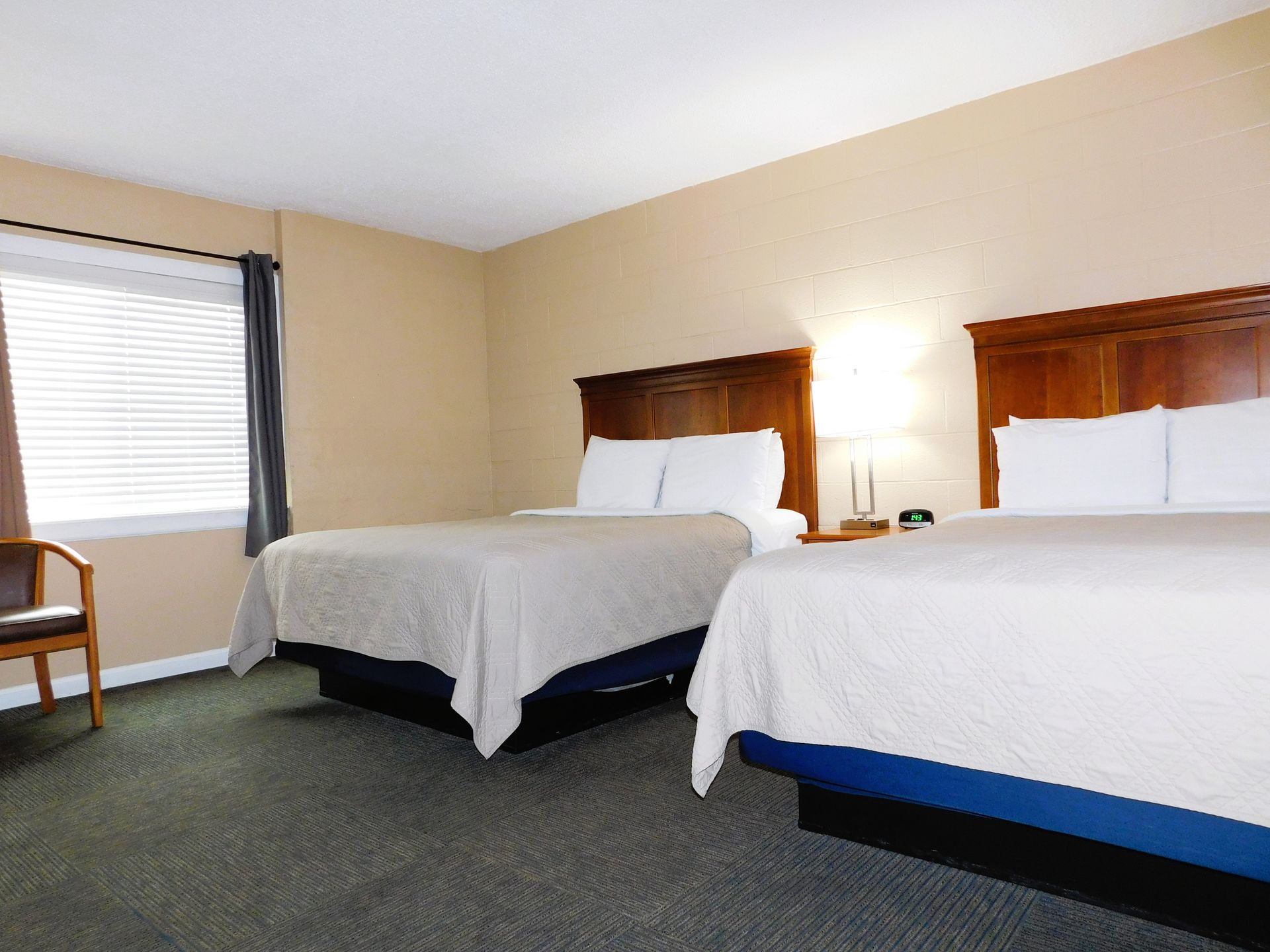 Hotel room with two queen beds, a window, and a chair. Beige walls, gray carpet.