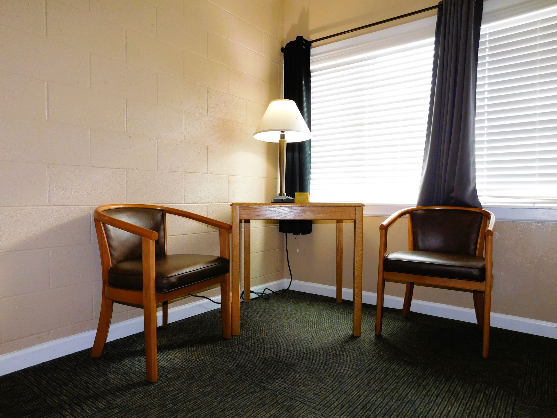 Two chairs, a table, and a lamp sit near a window with blinds, against a beige wall.
