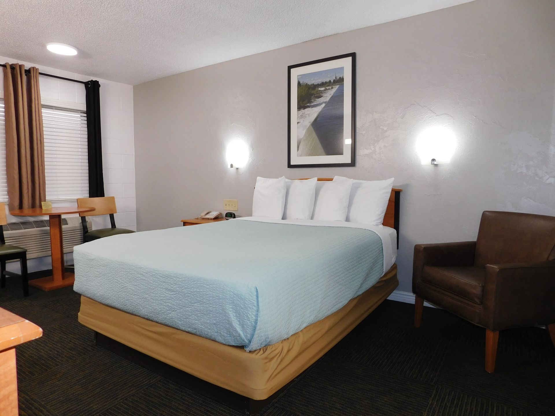 Motel room with a bed, armchair, table, and a framed picture on the wall.