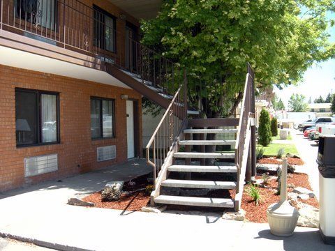 Exterior brick building with stairs leading up to an upper level, a tree, and parked vehicles.