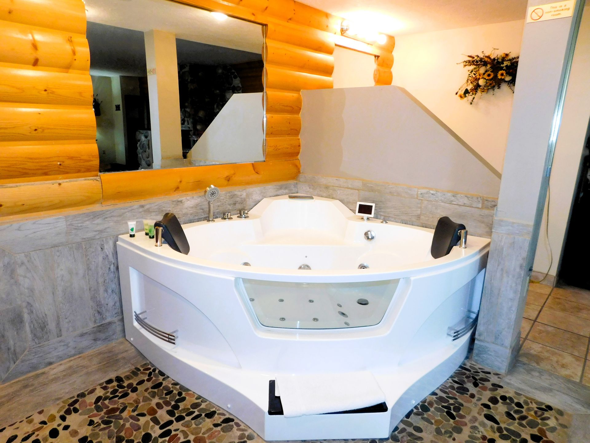 White corner jacuzzi in a room with wood paneling and stone flooring. Mirror above jacuzzi.