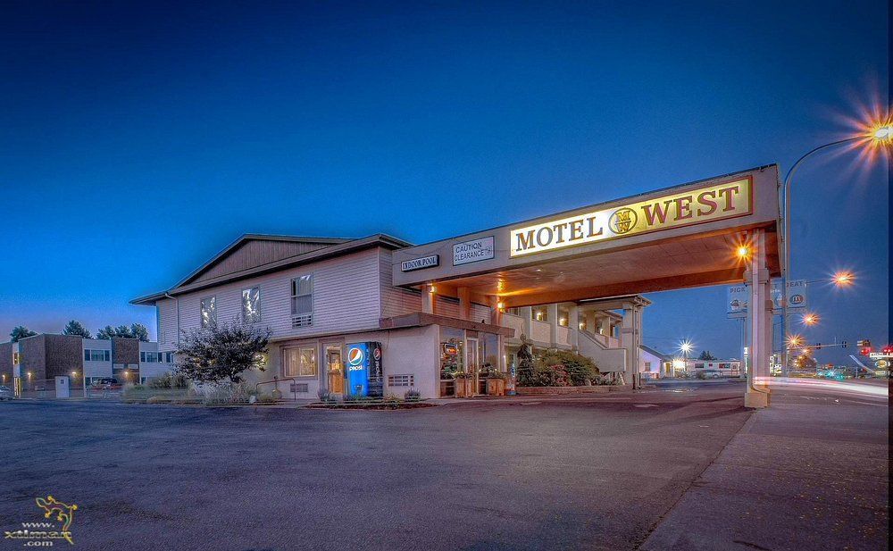 Motel West at dusk with a covered entrance, streetlights, and a blue sky.