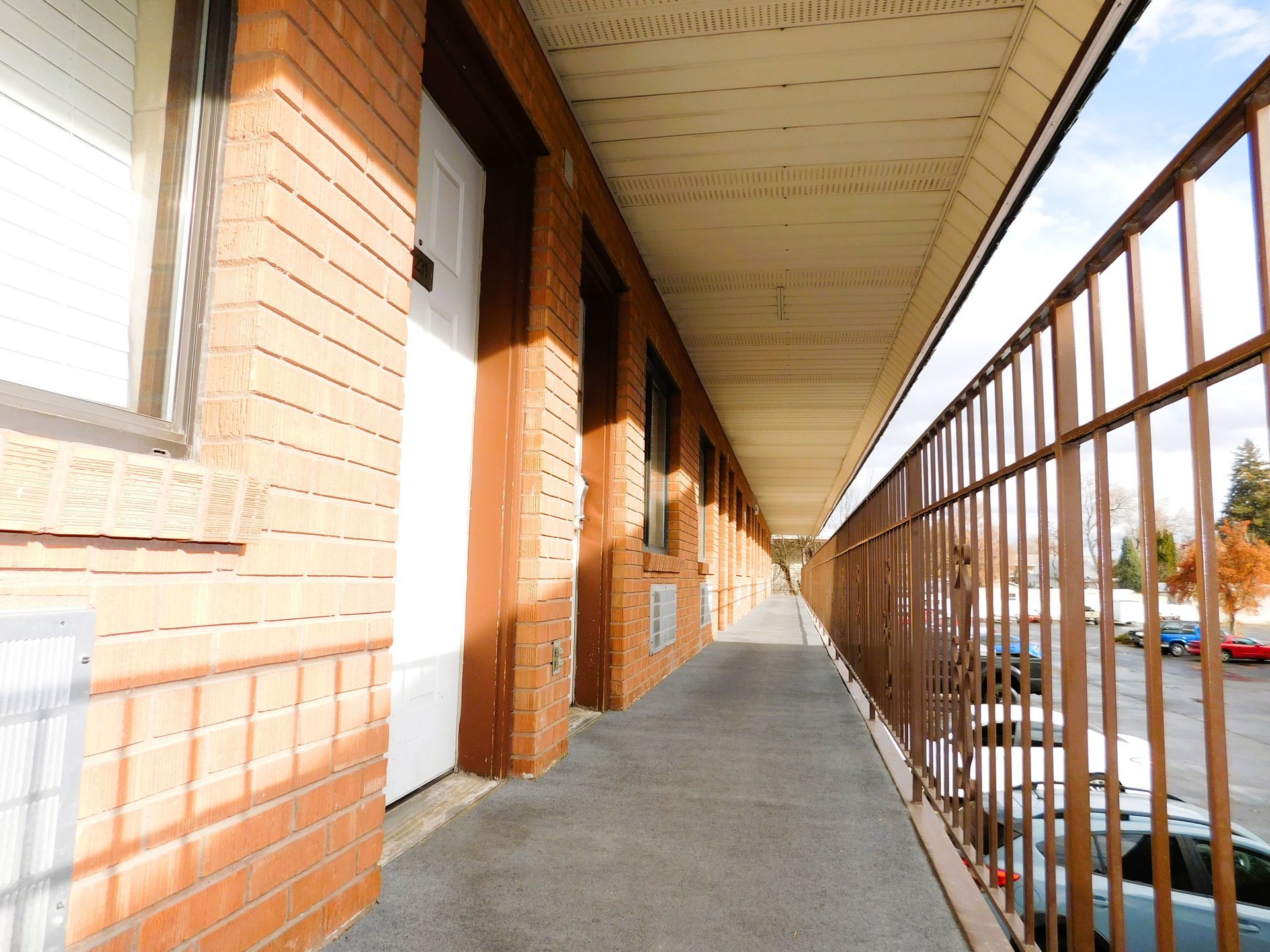 Exterior of a motel hallway with brick walls, doors, and a metal railing overlooking a parking lot.