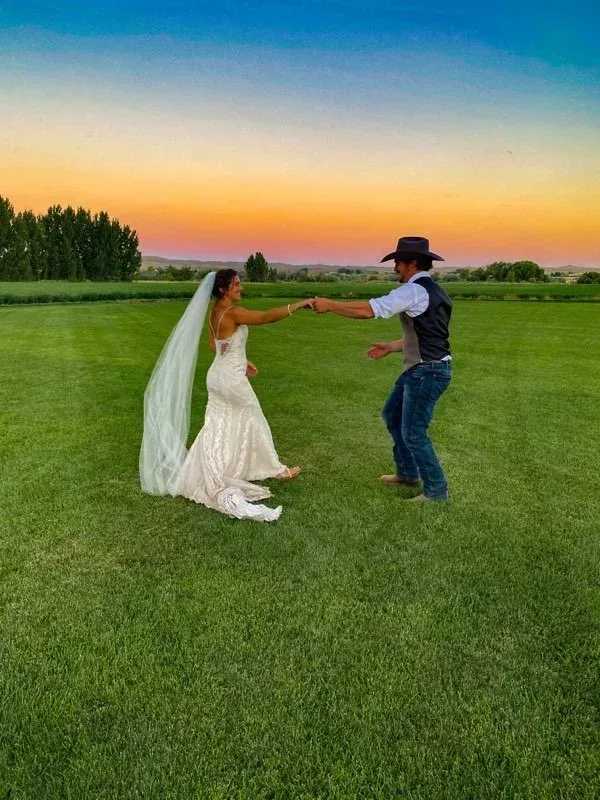 A couple in wedding attire dances in a grassy field at sunset, the bride’s long veil flowing behind her.