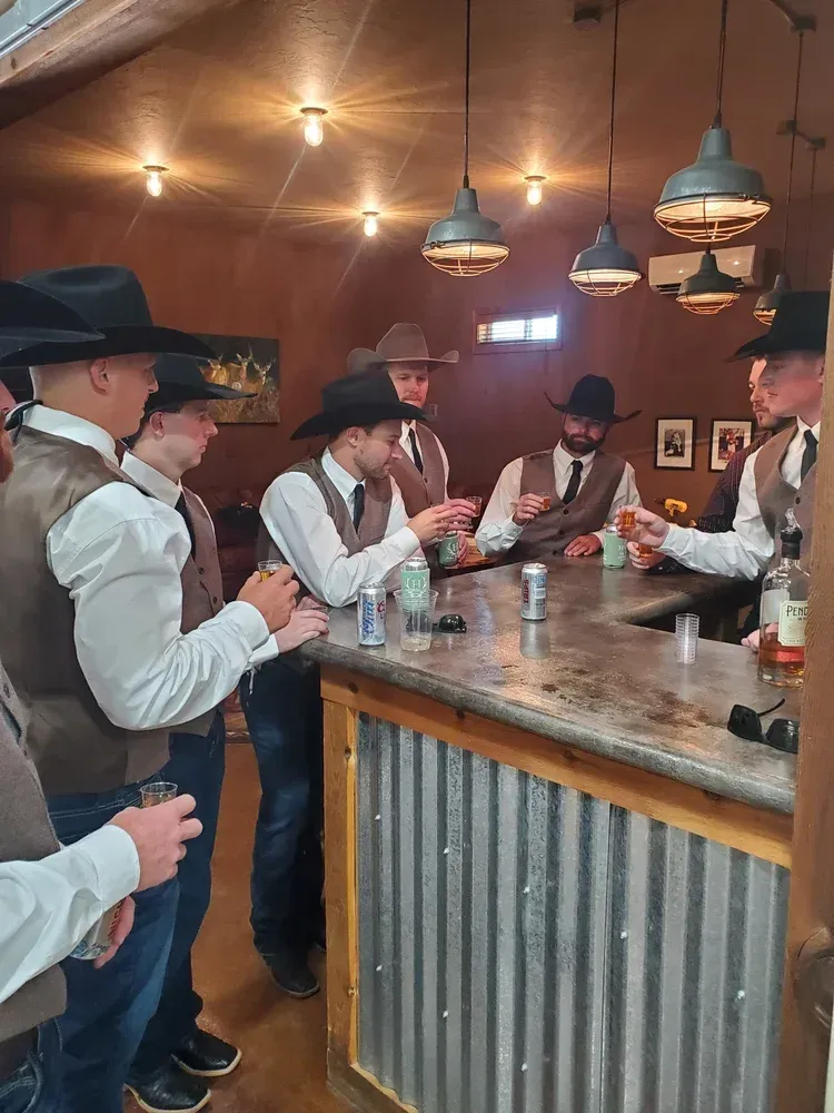 A group of people wearing cowboy hats and vests stand around a corrugated metal bar, holding drinks in a rustic room.