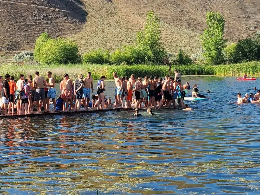 A large group stands on a wooden dock at a lake near hills, with several people swimming in the water nearby.