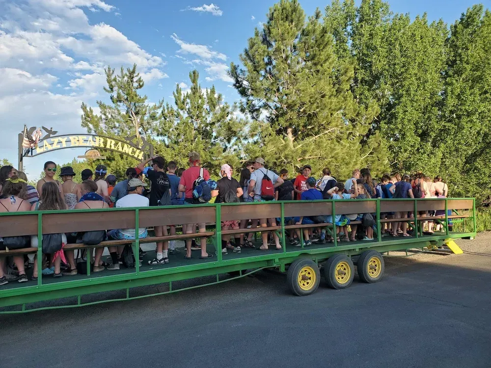 A large group of people sits on a green, multi-axle trailer passing under a sign for 