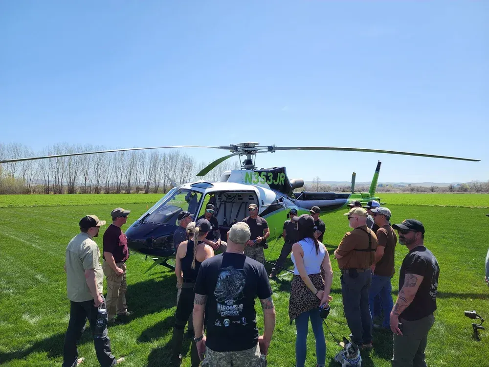 A group of people gathers in a grassy field around a white and green helicopter on a clear, sunny day.