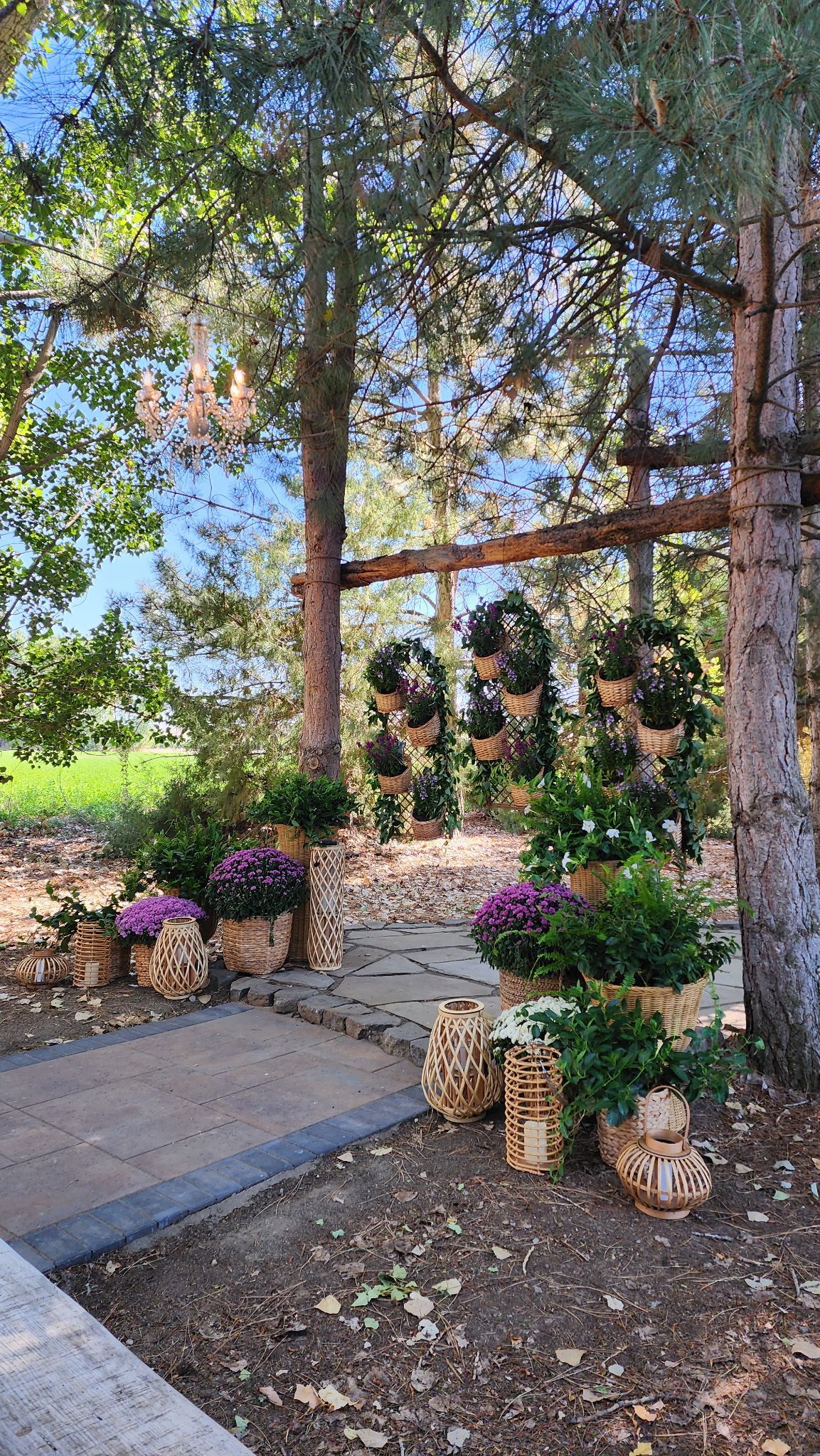 Outdoor wedding ceremony space with floral arrangements, hanging lanterns, and tree-lined stone path in a wooded setting.