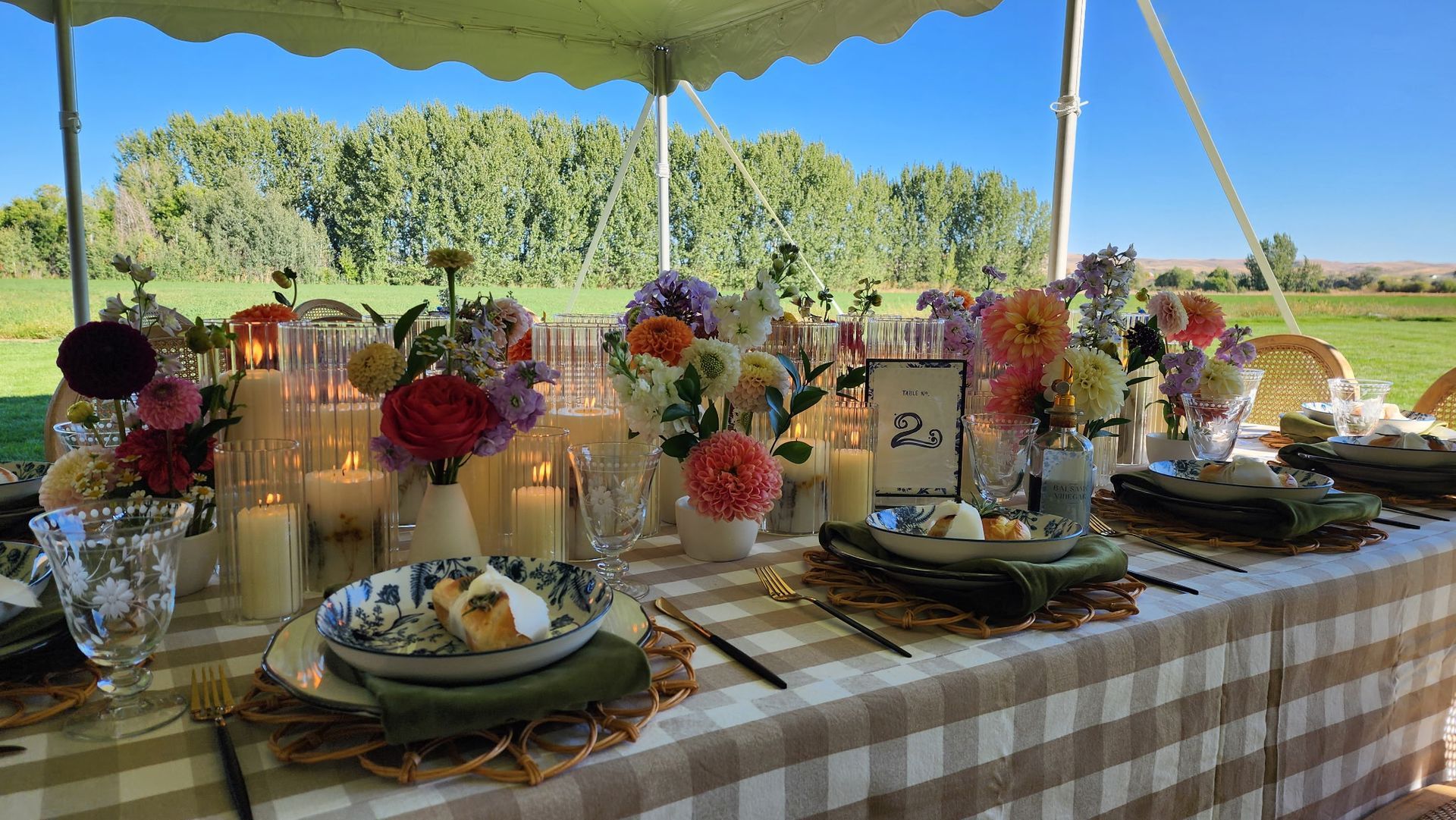 A long outdoor banquet table with a plaid tablecloth, decorated with vases of colorful flowers, candles, and place settings.