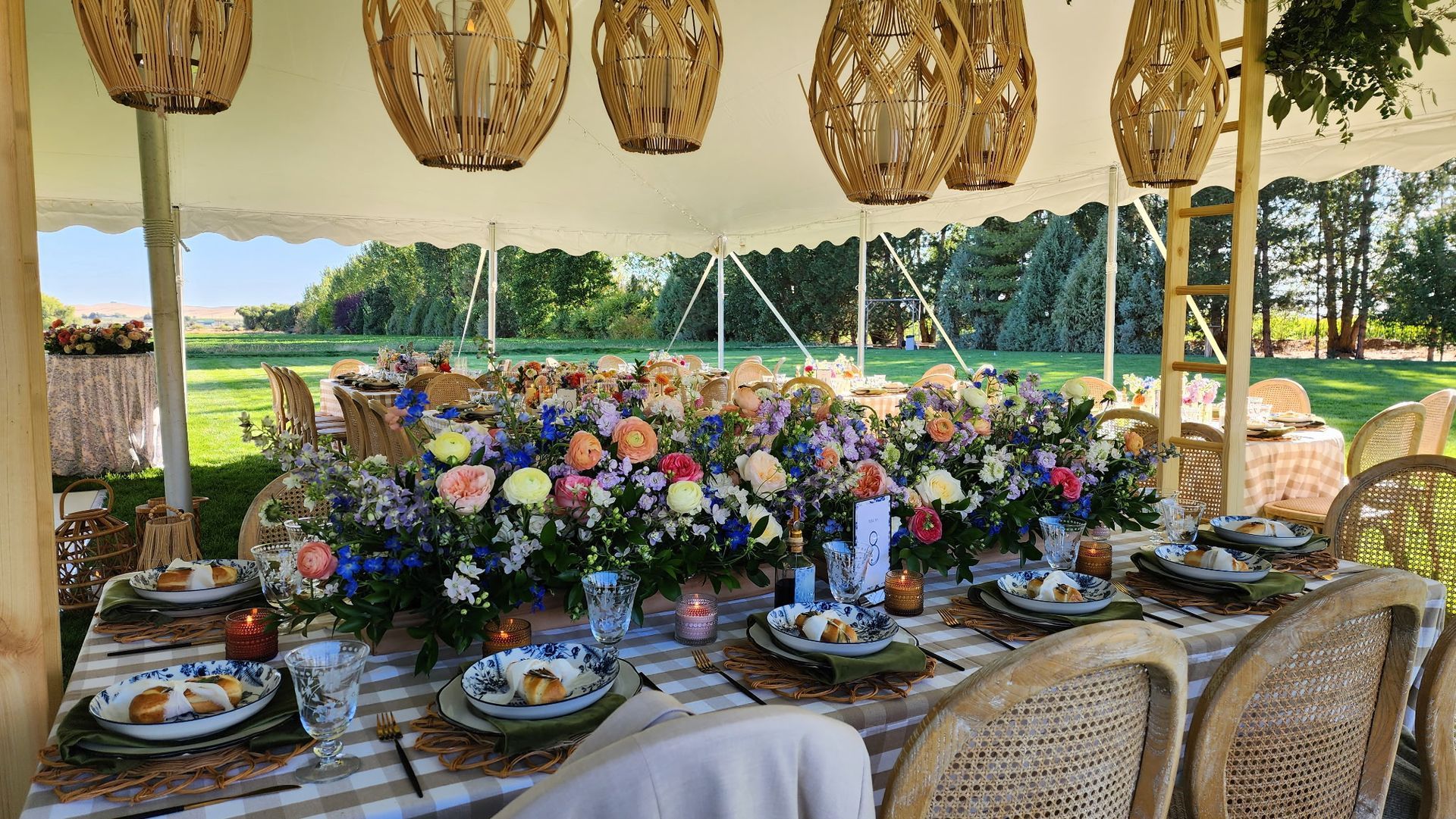 A wedding reception table under a tent, decorated with vibrant floral arrangements, hanging woven lamps, and place settings.