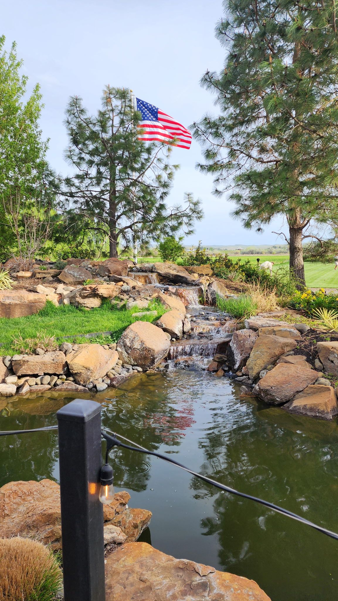 A multi-tiered waterfall flows into a tranquil pond surrounded by large rocks, lush green grass, and colorful flowers.
