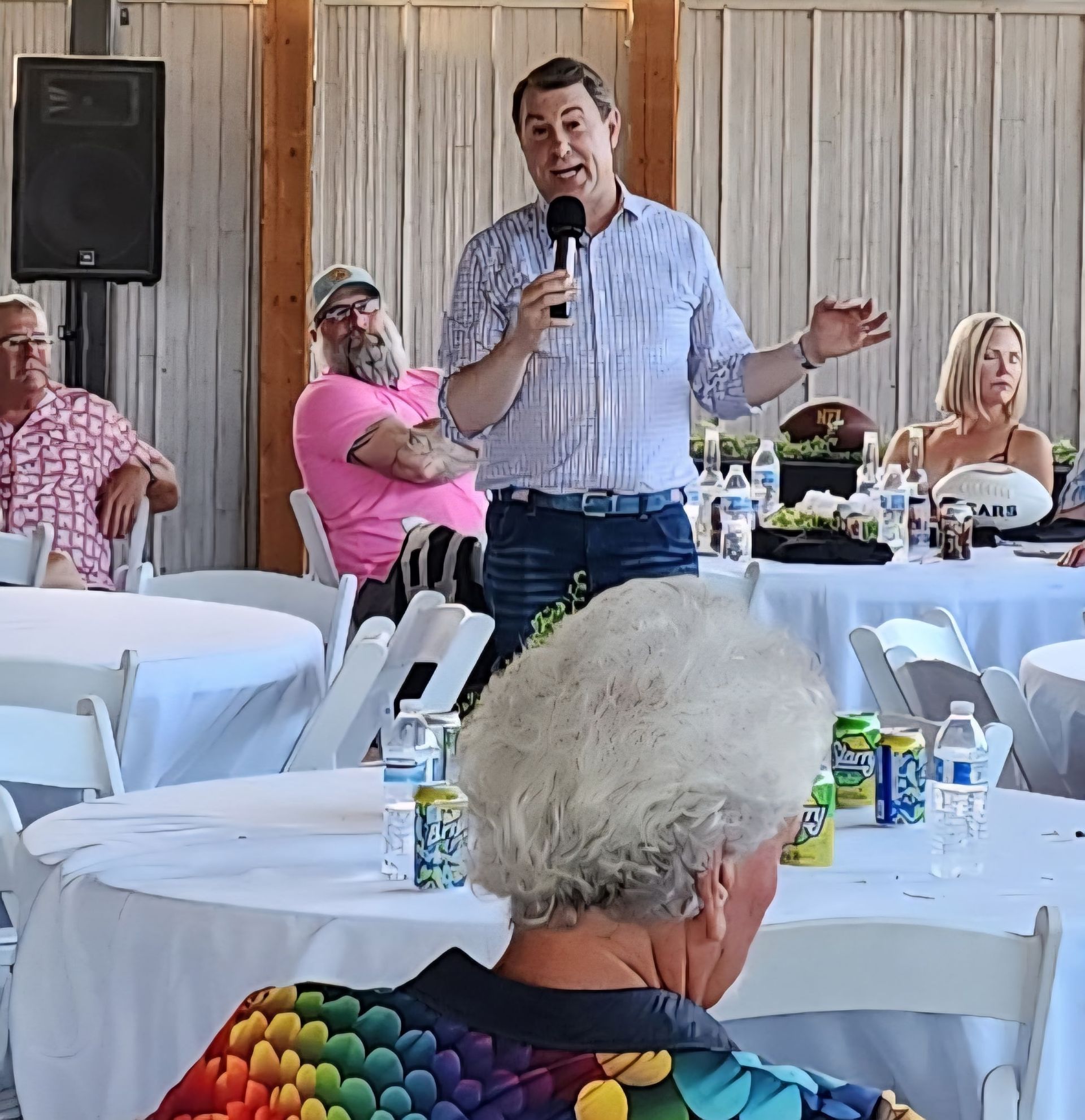 Man speaking into a microphone at a banquet hall, with seated guests and white-tablecloth tables around him