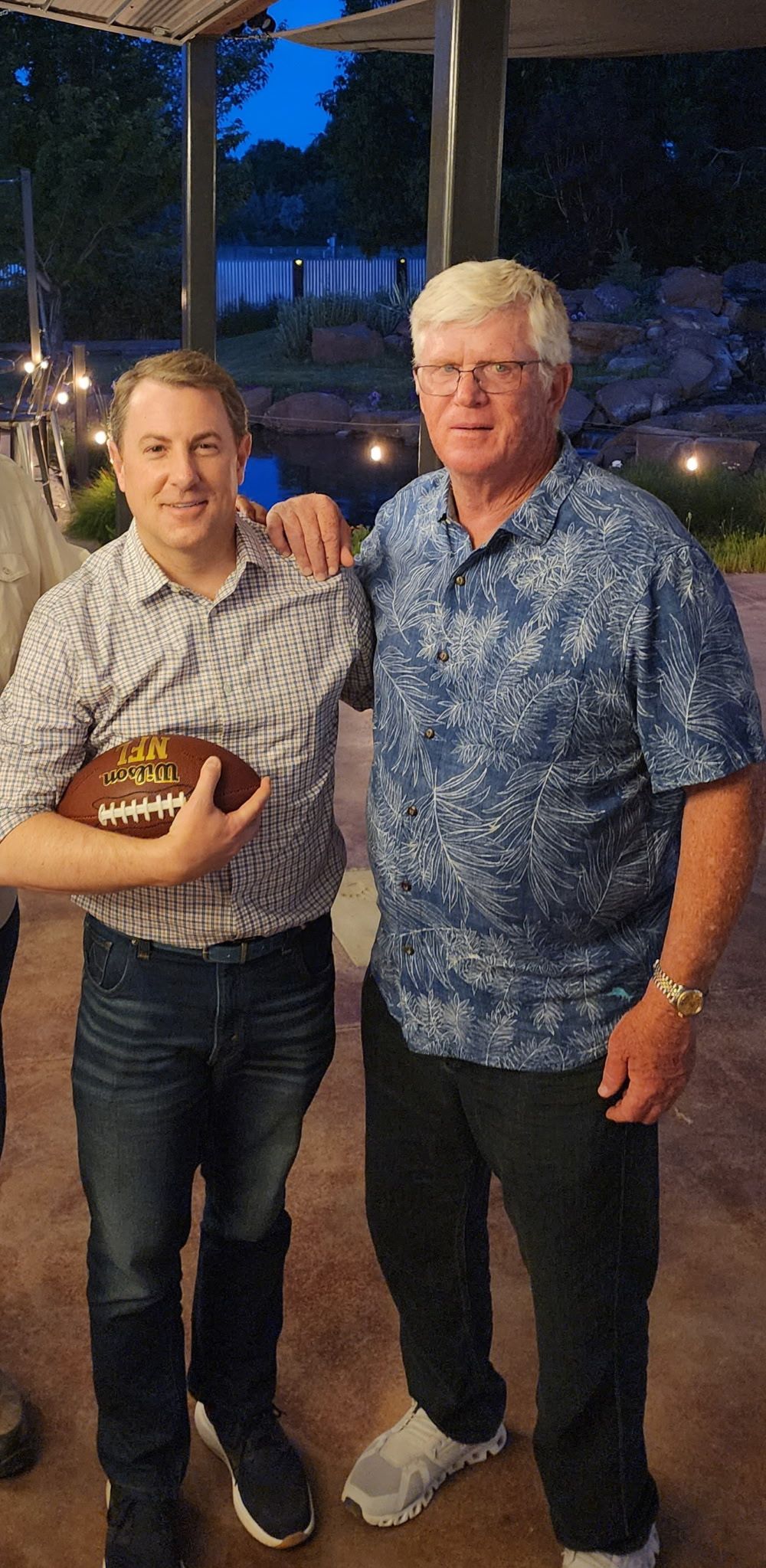 Two people standing outdoors at night, one holding a football, with lights and trees in the background.