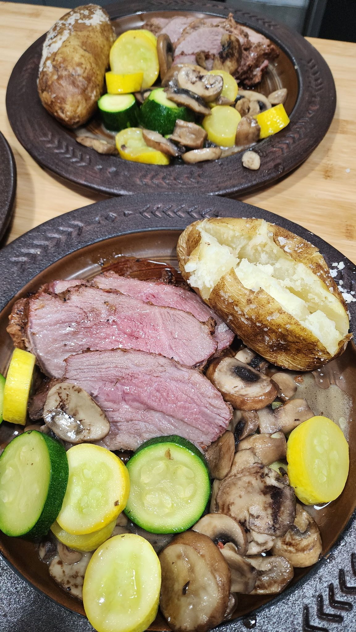 Two plates of steak, baked potato, mushrooms, and sliced squash on a wooden table