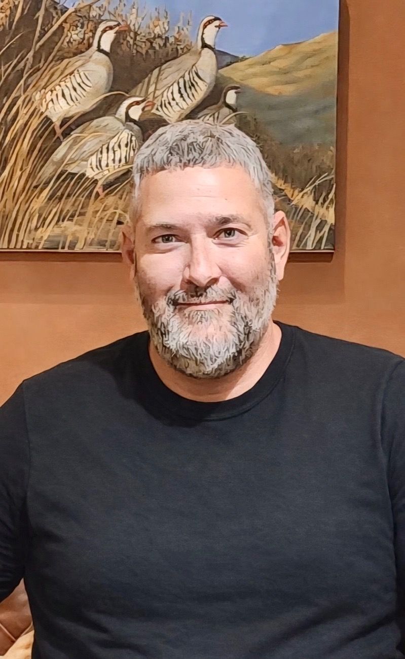 Smiling person in a black shirt standing indoors beneath a bird painting.