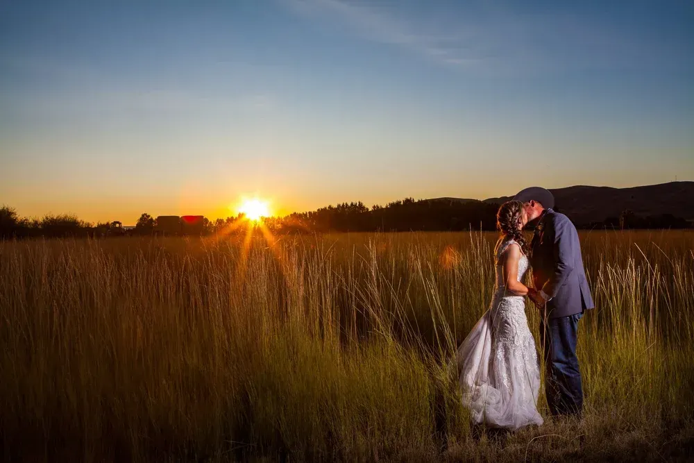 A couple in wedding attire embraces in a sunlit, grassy field at sunset.