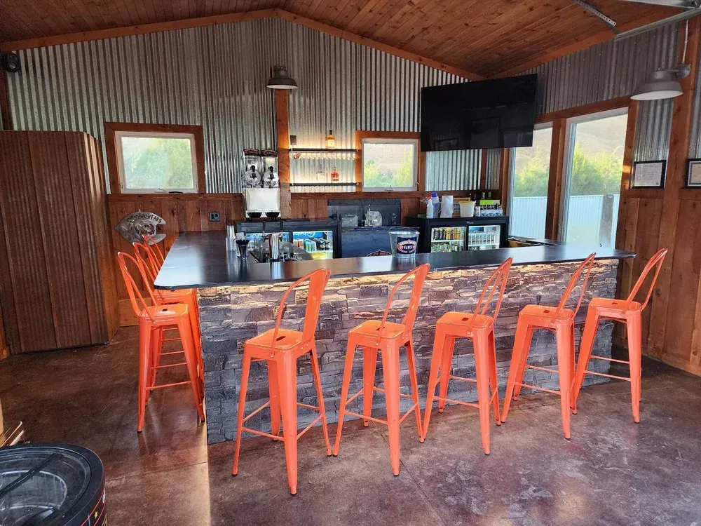 A rustic indoor bar with a stone-front counter, six orange metal stools, corrugated metal walls, and a mounted television.