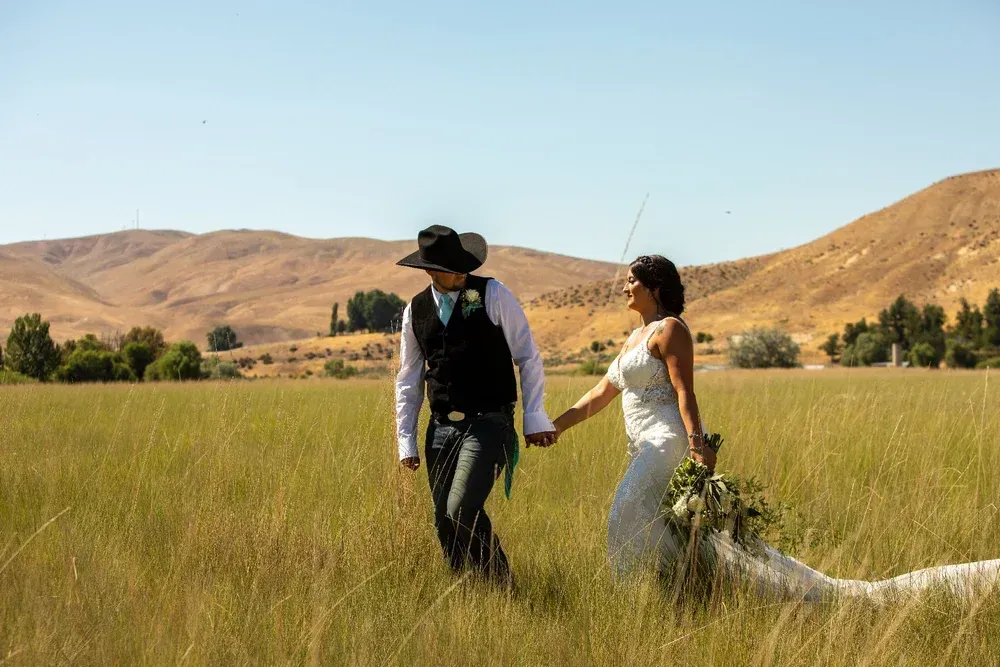 A couple in wedding attire walks hand-in-hand through a dry, grassy field against a backdrop of desert hills.