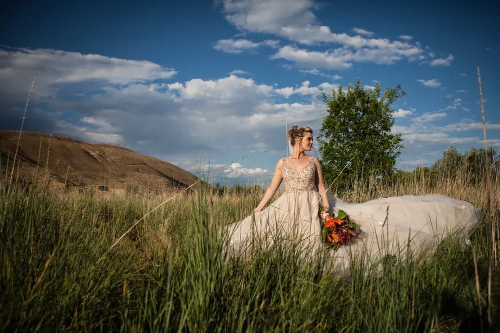 A person in a beaded wedding dress stands in a grassy field holding a bouquet, with a mountain and cloudy sky behind.
