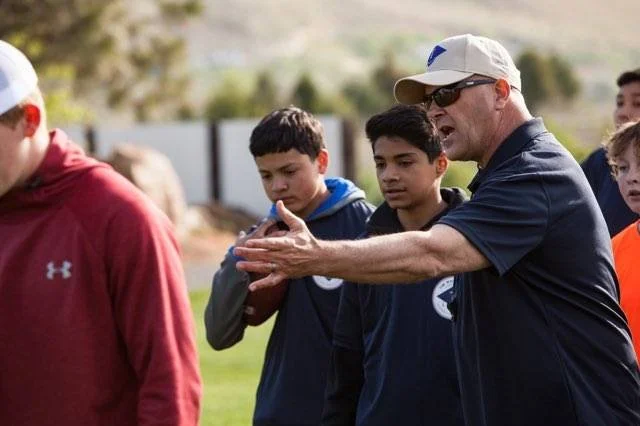 Coach in navy shirt and cap gesturing to two athletes during outdoor practice.
