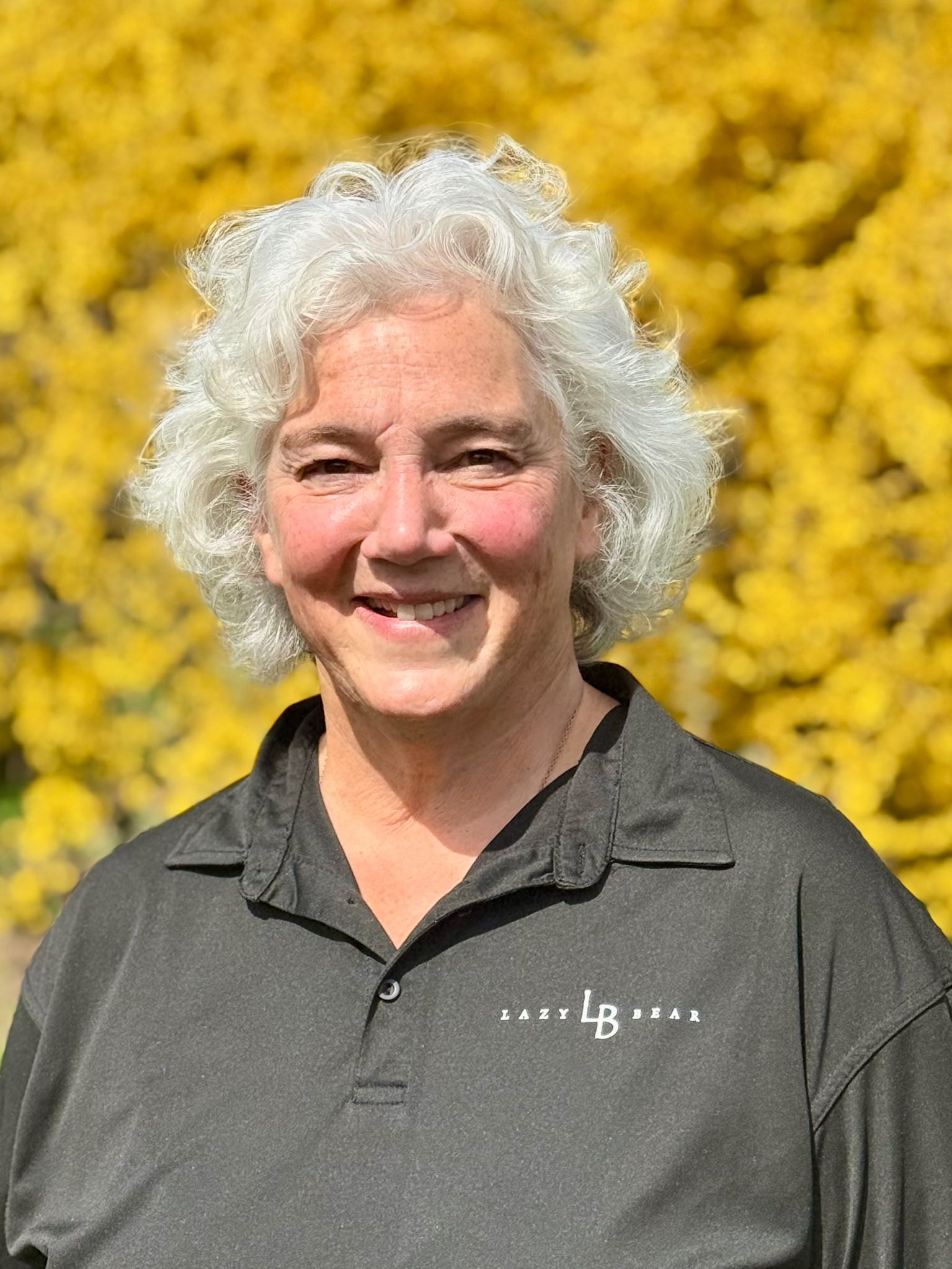Smiling person with curly white hair in a dark polo shirt, standing before bright yellow flowers