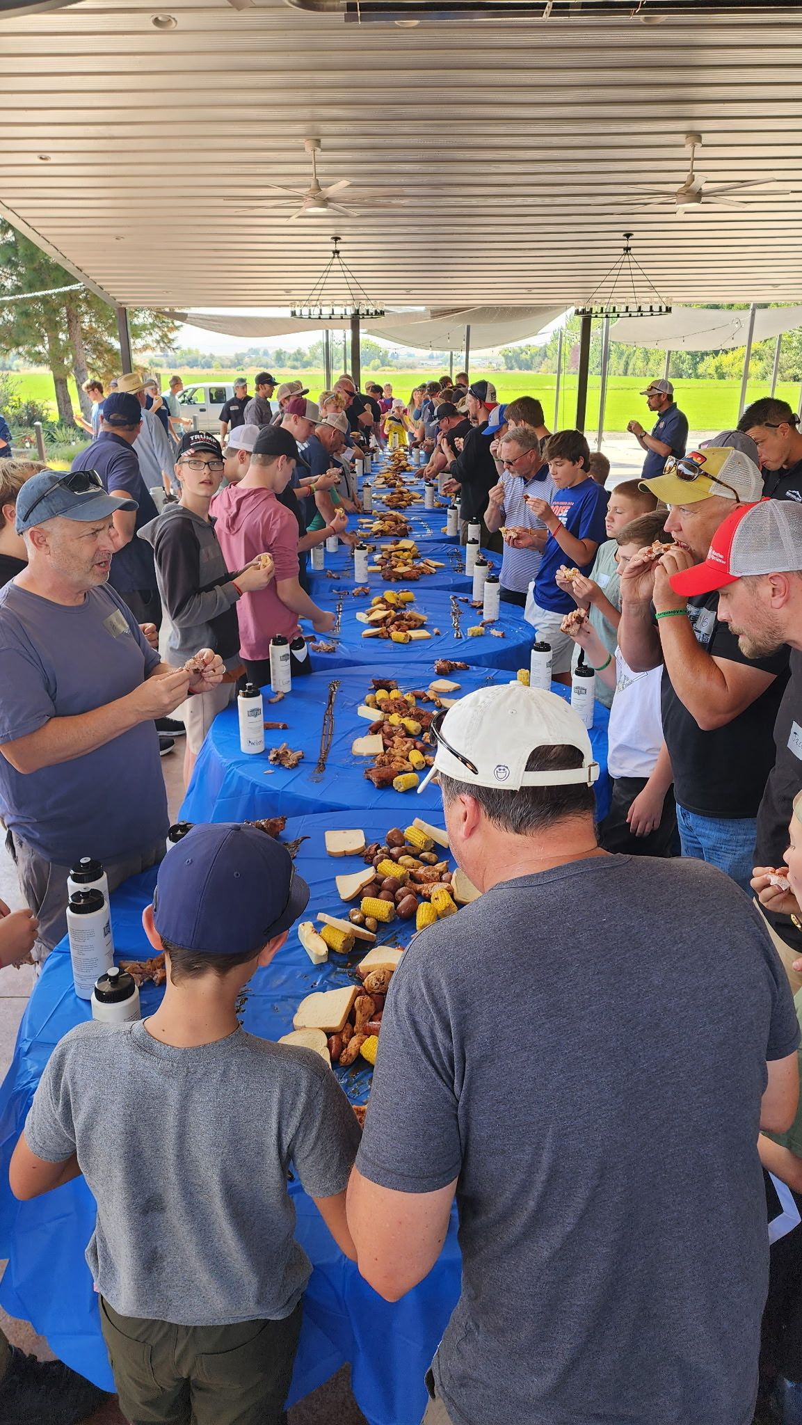 People gathered around long tables under a canopy, sharing a meal outdoors.