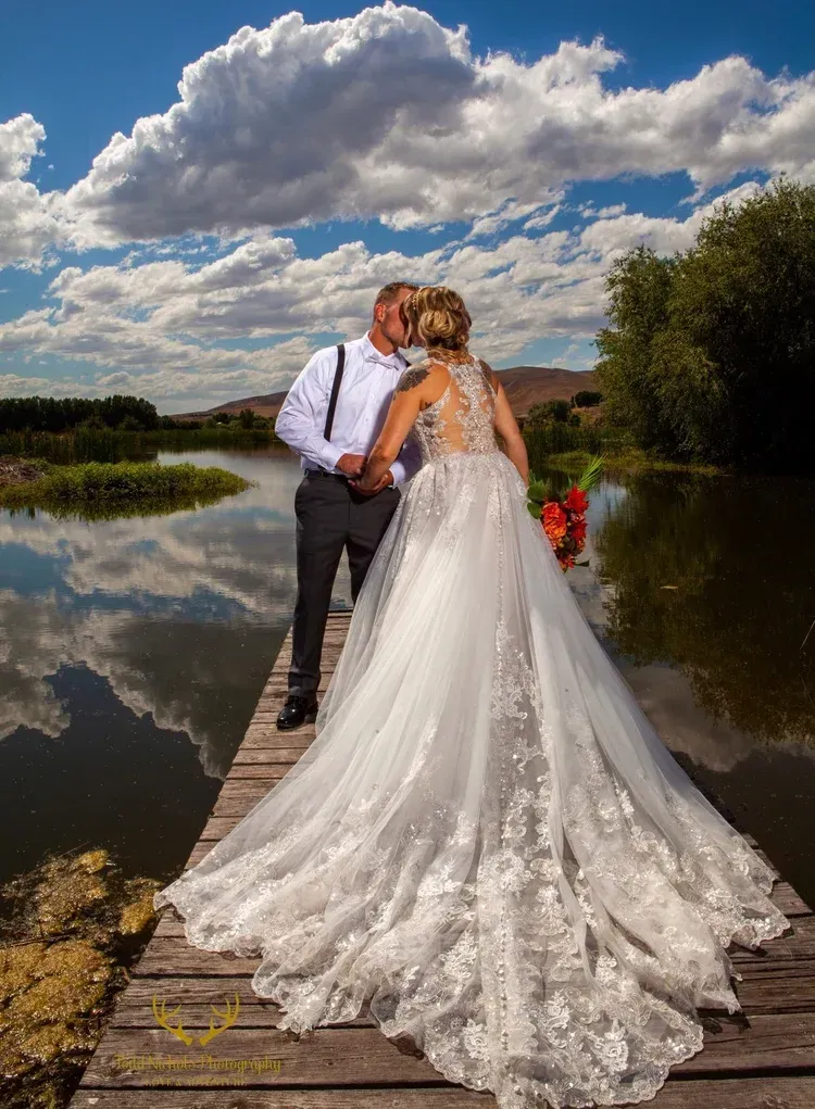 A bride and groom kiss in a sunlit field, the bride in a long white gown holding a vibrant orange and green bouquet.