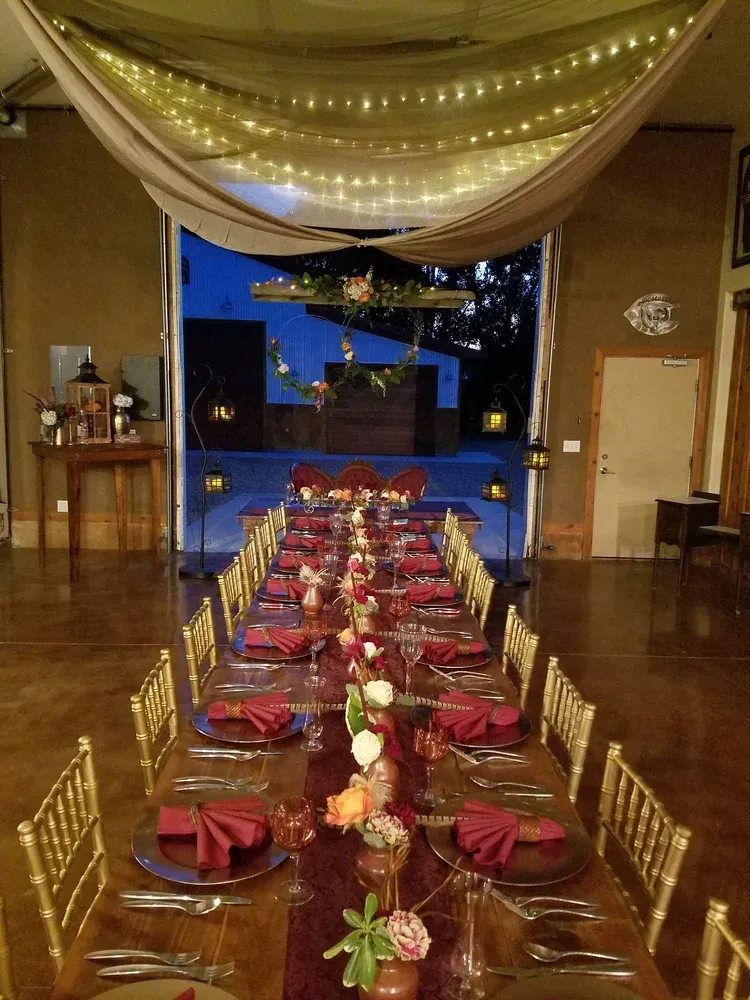 A long dining table set for a wedding or event with maroon napkins, gold chairs, and overhead draped lighting.