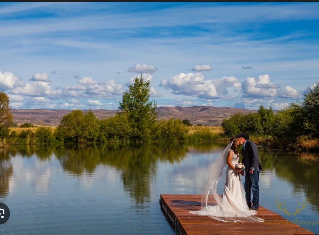A couple in wedding attire stands together on a wooden dock by a calm lake with trees and mountains in the background.