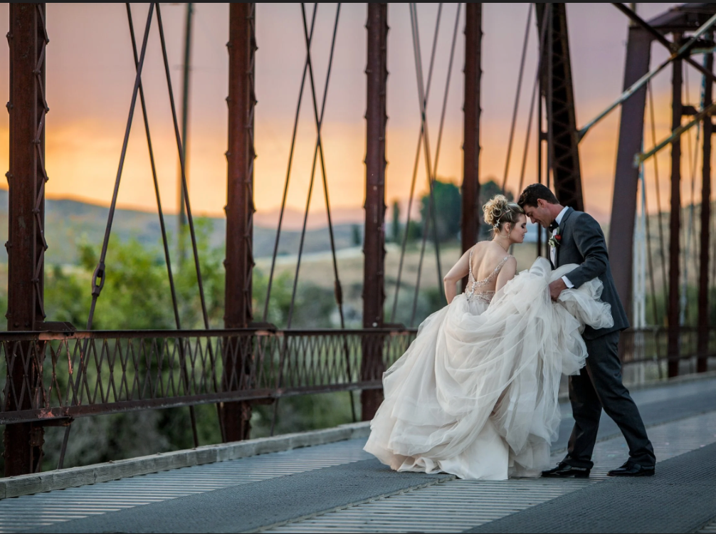 A couple in wedding attire walks across a rustic steel bridge at sunset, the person in the gown holding up its skirt.