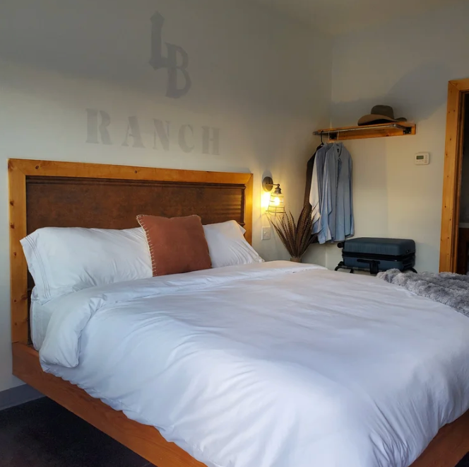 A modern, rustic-style hotel bedroom with a wooden bed frame, white bedding, and wall signage reading 