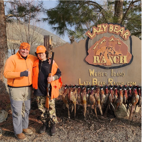 Two hunters in orange vests stand by a Lazy Bear Ranch sign in Weiser, Idaho, with a row of harvested pheasants in front.