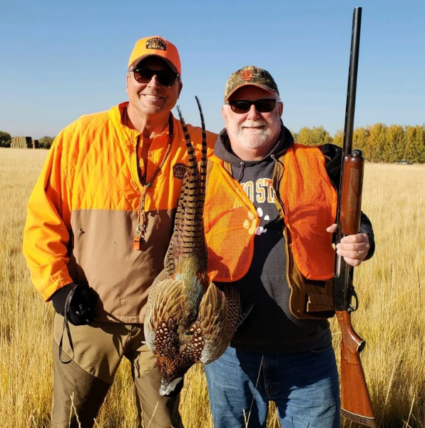 Two hunters in orange vests stand in a field, one holding a pheasant. Both wear sunglasses and hats under a blue sky.