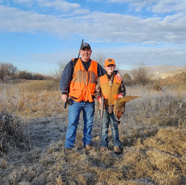 Two hunters in blaze orange vests standing in a field with a bird, holding shotguns under a blue sky.