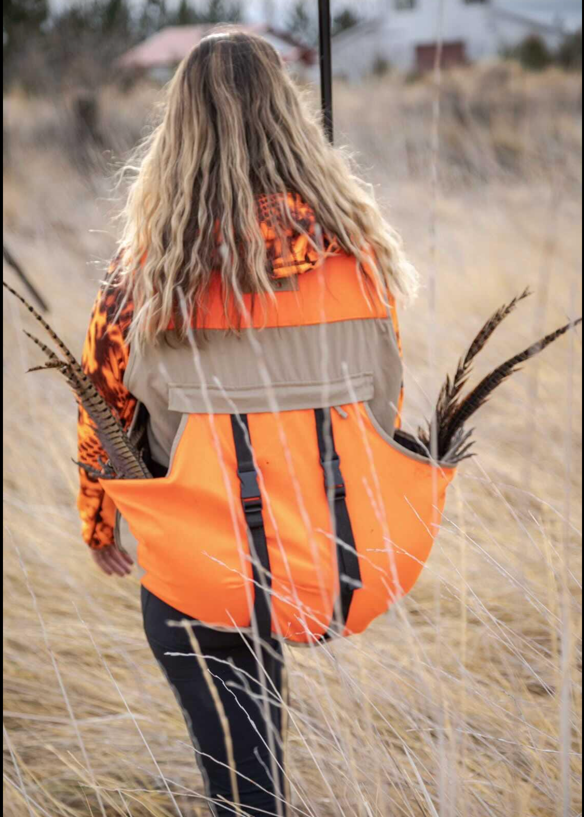 A hunter wears a blaze orange vest with a game bag containing pheasant feathers in a grassy field.