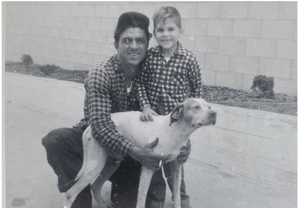 A person and a child wearing plaid shirts pose kneeling with a light-colored dog against a block wall.