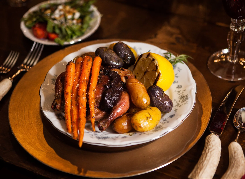 A roasted chicken dinner served with glazed carrots, multi-colored potatoes, and a lemon wedge on a formal dinner table.