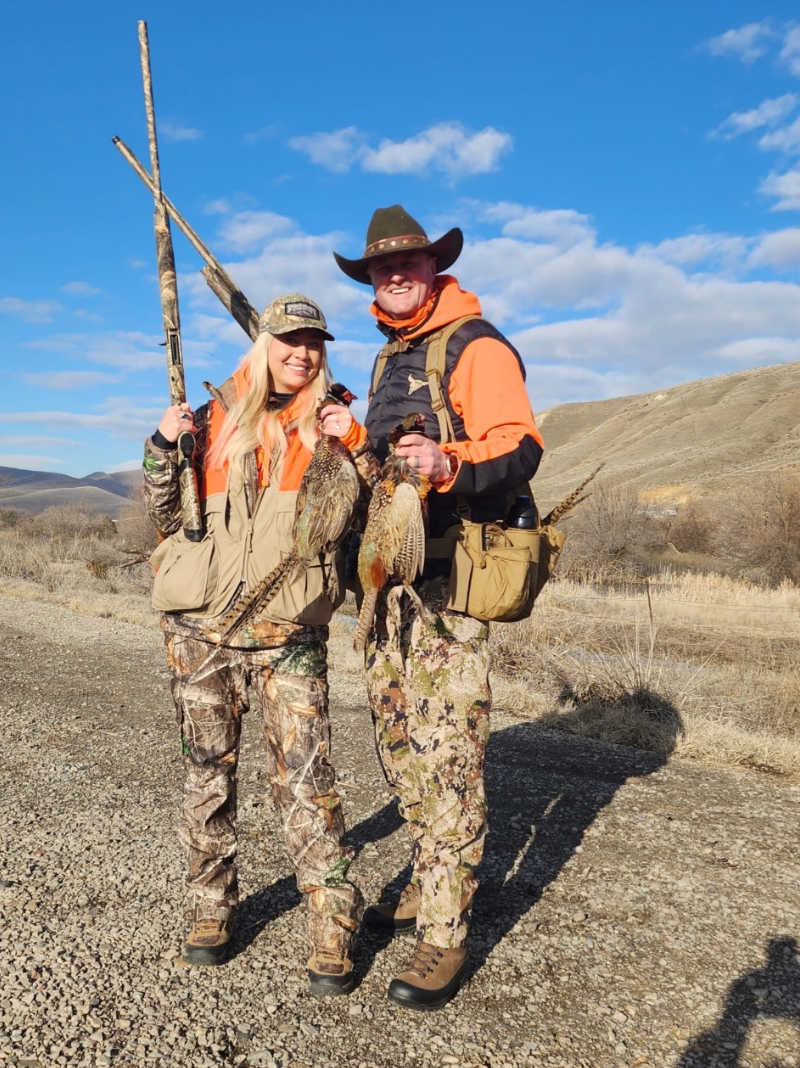 Two hunters in camouflage and orange vests pose outdoors on a sunny day, holding shotguns and harvested game birds.