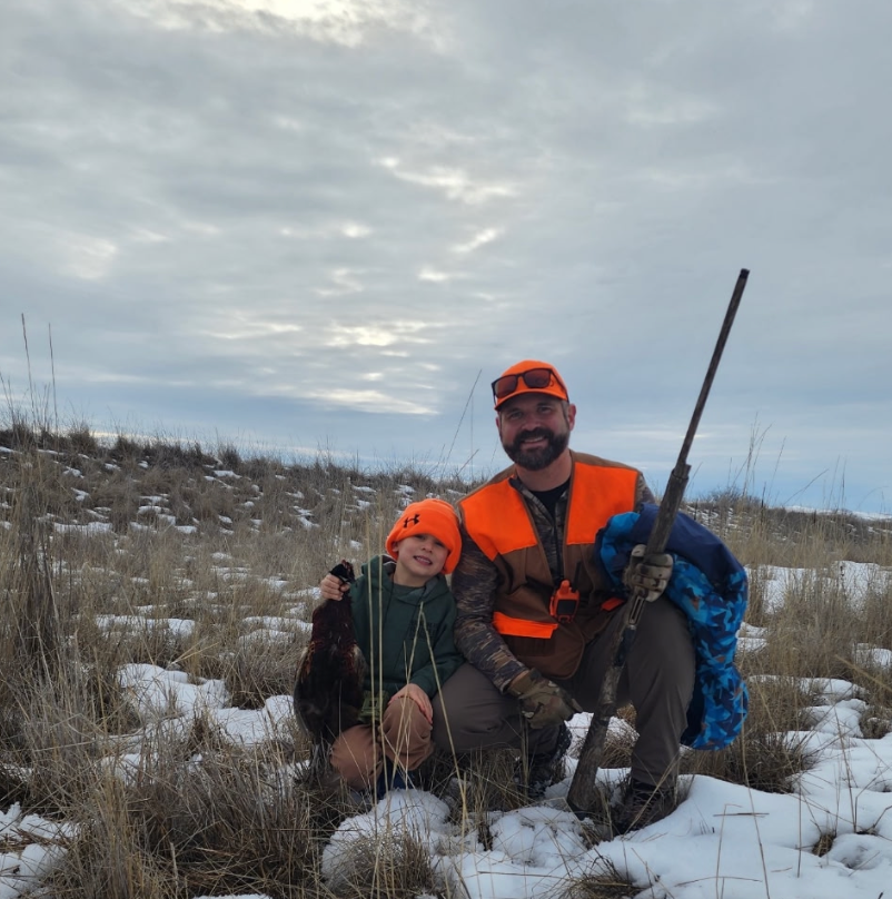 A person and a child in bright orange hunting hats and vests crouch in a snowy field, holding a harvested bird and a gun.