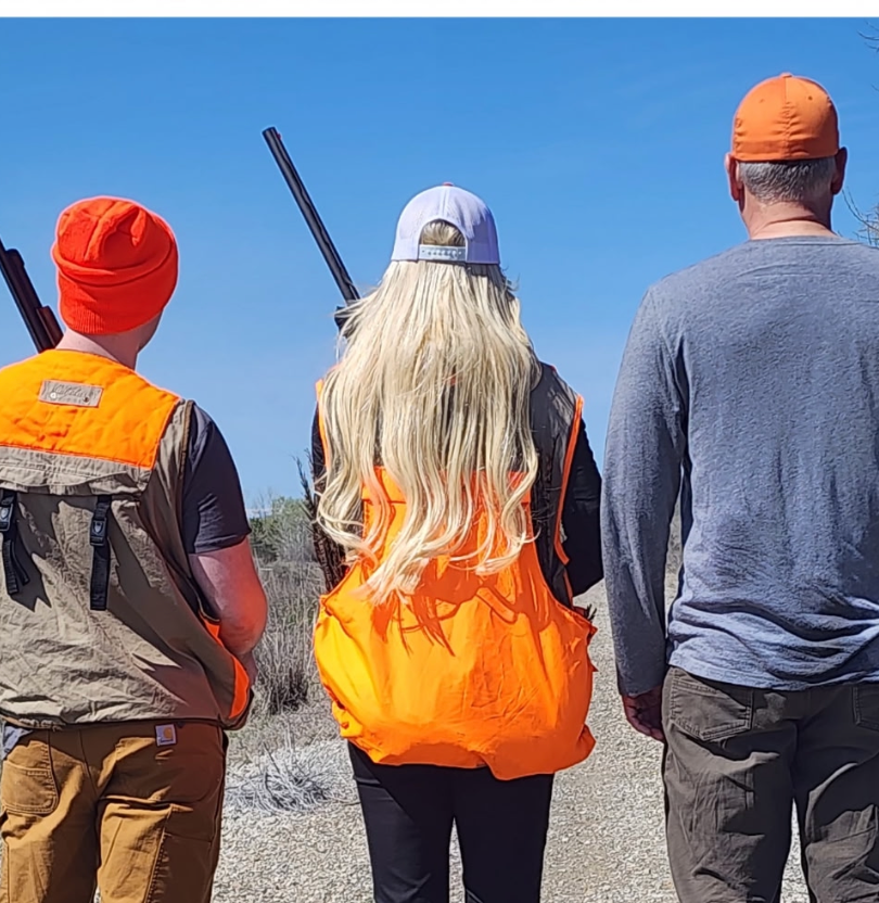 Three people in high-visibility orange hunting gear and hats stand outdoors, holding rifles and looking toward the horizon.