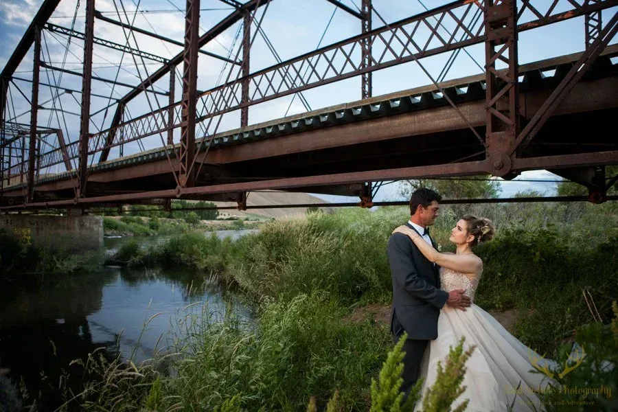 A bride and groom embrace in a meadow under a large metal bridge next to a flowing river.