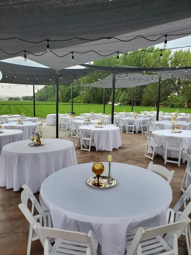 Outdoor event space with white-clothed round tables, white chairs, and string lights under a grey canopy.