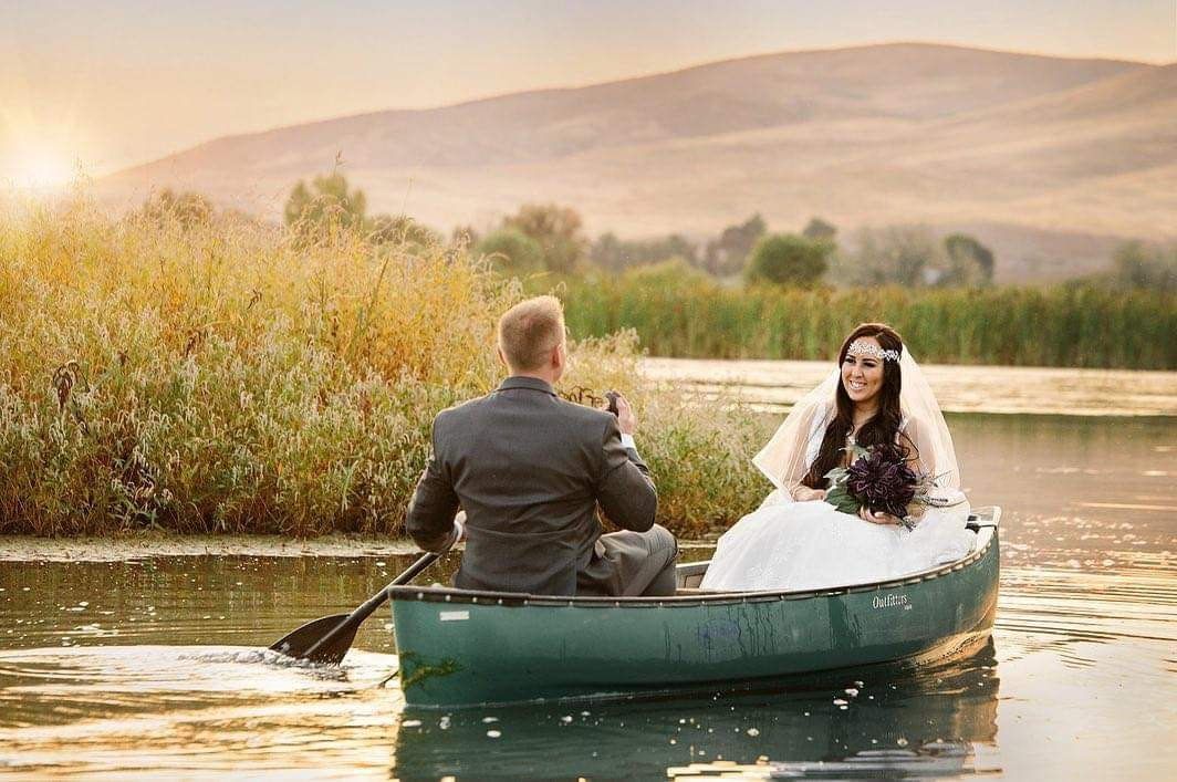 A person in a suit paddles a canoe carrying a person in a white wedding gown on a calm lake at sunset.