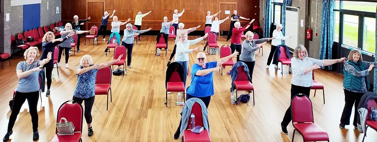 People enjoying a gentle keep fit class standing behind their chairs while smiling and swinging their arms.