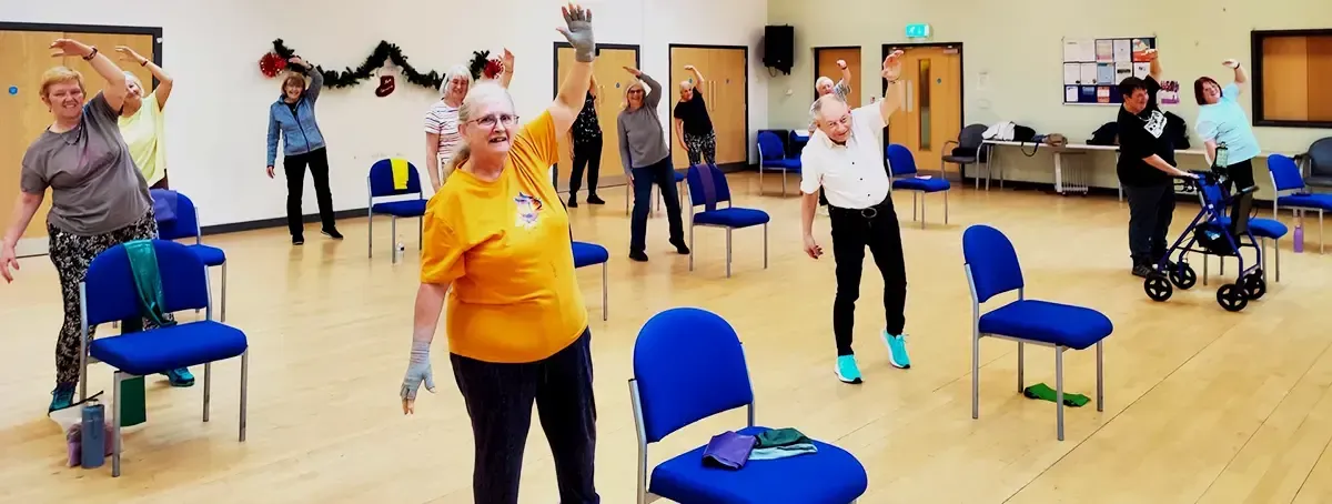 People in a gentle keep fit class in a community centre smiling while stretching one arm over their heads.