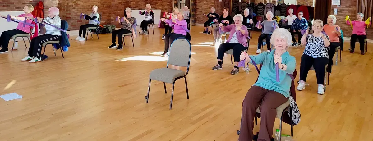 People seated in a gentle keep fit class doing resistance band exercises to strengthen their upper body.